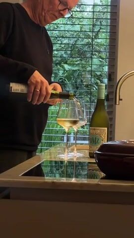 A person is pouring white wine from a bottle into two wine glasses. The wine glasses are tall and clear, and they are placed on a reflective surface. There is another bottle of wine next to the glasses, and it is also clear with a label that says "DMZ". The background is out of focus, but it appears to be a window with blinds, and outside there are trees and greenery. The lighting is dim and moody.