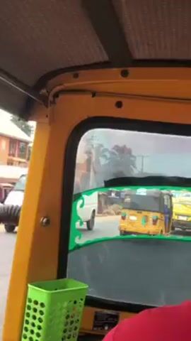 A first-person perspective video shows the view from inside a yellow tuk-tuk. The driver is a dark-skinned man wearing a red shirt. Through the back window, another yellow tuk-tuk is visible, followed by yet another. The road appears to be paved. A green plastic basket hangs inside the tuk-tuk, on the left. The interior of the tuk-tuk is mainly yellow with a patterned gray roof. Green decorative trim adorns the back window frame. The scene seems to be taking place in an urban environment.