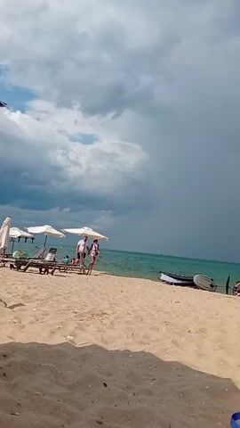 A beach scene with several people. The sky is cloudy and overcast. The sand is light brown. There are several beach umbrellas and lounge chairs visible. Two people are walking near the water's edge. A small boat with an outboard motor is on the sand near the water. One person, wearing black shorts and a bikini top, is standing and bending over near a lounge chair. Another person is lying on a lounge chair covered with a blue towel. The camera is positioned low, giving a view of the sand and the legs of a person in the foreground.