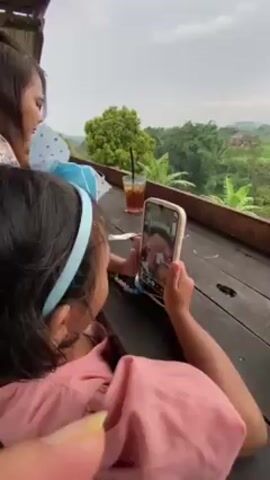 Here's a description of the video:

The video shows a young girl sitting at a wooden table, engaged in a video call on a smartphone. She is wearing a pink top and a light blue headband. The phone is positioned to show both her and the person on the other end of the video call. In the background, there's a view of lush greenery with trees and possibly distant houses. There's also a glass of iced tea on the table and another person can be seen in the background holding up another phone, potentially taking a photo or video. The little girl turns and smiles towards the camera.