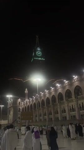 The video shows a night view of a plaza with people gathered. In the background, a large clock tower is illuminated with green lights. The tower has Arabic inscriptions. In front of the tower, a building with arched windows and many lights is visible. People are seen walking in the plaza, most of them wearing white garments. A lamppost with bright lights is in the foreground. The overall scene suggests a gathering at a religious or historical site at night.