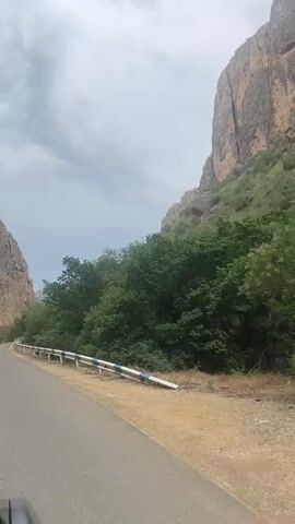 The video presents a first-person view from a vehicle traveling along a winding paved road. A white and blue guardrail lines the right side of the road. On both sides, large, steep, light brown rocky cliffs rise sharply, with dense green trees and bushes covering their lower sections. The sky above is overcast with gray clouds. The camera maintains a forward perspective, with a slight upward pan at the end focusing on the cliff face to the right.