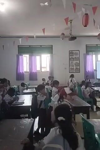 A classroom scene with approximately fifteen students, mostly young children, seated at desks. The students are wearing school uniforms, some of which appear to be scout uniforms with sashes. The classroom is decorated with red and white triangular flags and two paper lanterns hanging from the ceiling. There are windows with purple curtains, a projector mounted on the ceiling, and a whiteboard or screen at the front of the room. Some students are looking forward, possibly at a teacher or screen, while others are looking down at their desks or interacting with each other. One student in the foreground is looking towards the camera. The overall atmosphere appears to be a school lesson or activity.