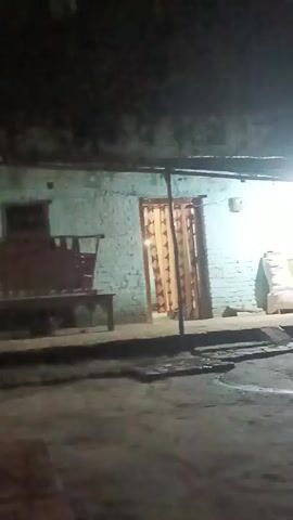 A poorly lit outdoor scene at night. A light illuminates a simple building made of light blue brick. A covered porch extends from the building, supported by poles. There is a door with a curtain visible behind it. To the right of the door, there are several sacks, one white and others yellowish, stacked against the wall, along with some other objects. A window with a wooden frame is visible to the right of the sacks. In the foreground, the ground appears to be dirt or gravel, with some areas wet or reflective, suggesting puddles. To the left of the building, a dark object that could be a bench or furniture is partially visible. A tree with dark foliage is located to the right of the building, obscuring part of the scene. The overall lighting is dim, with a distinct light source directly above the porch area.
