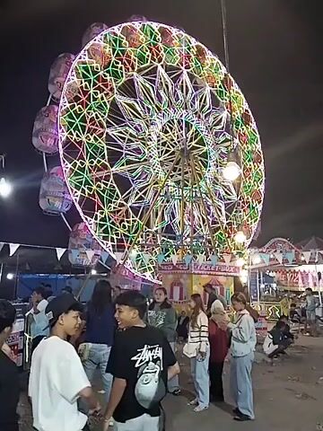 The video shows a bustling night scene at what appears to be a carnival or night market in Sragen, Indonesia. The dominant feature is a large Ferris wheel, brilliantly illuminated with intricate patterns of colorful LED lights in shades of green, red, yellow, and purple. The Ferris wheel has multiple gondolas, some of which are also lit.

Below the Ferris wheel, a crowd of people, mostly young adults, are gathered. They are dressed in casual attire, including t-shirts, hoodies, and jeans. Many are seen chatting, looking around, and interacting with each other. Some individuals are facing the camera, while others have their backs turned or are looking towards the rides and stalls.

In the background, other amusement park structures and booths are visible, also adorned with lights. One booth clearly displays the text "TICKET BOX," indicating it's where tickets for attractions are sold. The sky is dark, highlighting the vibrant artificial lighting of the carnival. The atmosphere appears lively and festive, with people enjoying the evening out. The ground is unpaved, suggesting a temporary setup for the event.