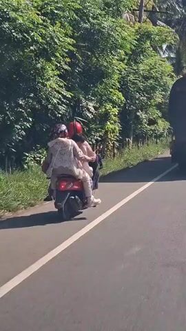 Two people are riding on a red scooter on a paved road. The person in the front is wearing a red helmet and a light brown jacket. The person in the back is wearing a white patterned jacket and a red helmet. The scooter is moving forward on the right side of the road, which has a white line marking the lane. The road is lined with green trees and vegetation. A portion of a large dark vehicle is visible on the right side of the frame, suggesting the scooter is overtaking it or passing it. The sky is visible above the trees, appearing bright and possibly sunny. The overall scene depicts a road trip or travel through a rural or tropical environment.
