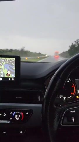 The video shows an interior view from a car, looking out the front windshield during heavy rain. The windshield wiper repeatedly sweeps across the wet glass. Ahead, on a multi-lane wet road, a large red truck with its tail lights illuminated is visible in the distance, followed by other vehicle lights. The car's dashboard features a black steering wheel with an Audi logo, a digital speedometer displaying "143," and multiple illuminated warning lights including check engine and tire pressure. A central infotainment screen on the left displays a navigation map with a blue marker and a "143" indicator. Below the screen, a climate control panel shows "21.0." Trees and greenery line the sides of the road.