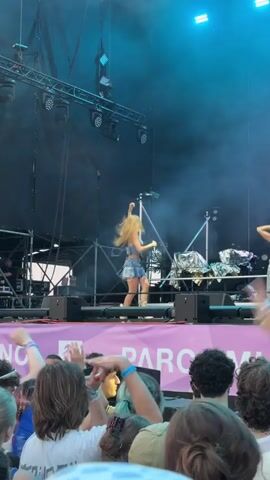A live music performance is taking place on a stage under bright stage lights and a smoky atmosphere. A female singer with long blonde hair, wearing a blue and white patterned top and a denim skirt, is performing center stage, holding a microphone. To her right, two other performers, one female and one male, are also on stage, dancing. The stage is set up with speakers and various equipment, including what appears to be large silver foil-covered objects. In the foreground, a crowd of people are visible, many with their arms raised in the air, enjoying the performance. The backdrop of the stage features a large screen displaying a sparkling blue pattern. A banner with purple lettering is visible across the front of the stage, partially obscured by the crowd.
