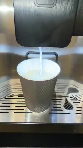 A close-up, medium shot captures a coffee machine dispensing a creamy liquid into a gray and white paper cup. The cup sits on a stainless steel drip tray with horizontal slats and teardrop-shaped drainage holes. The liquid stream is white and frothy, creating ripples as it fills the cup, which is about halfway full. The background shows the black front of the coffee machine, with a glimpse of a white illuminated panel at the top. The overall setting appears to be indoors, likely a cafe or kitchen. The action is focused entirely on the dispensing of the beverage.