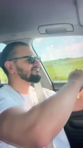 The video shows a man driving a car on a sunny day. The man has a beard and is wearing sunglasses and a white t-shirt with red text that appears to say "CHRISTIAN ROOTS 1945-1950". He is smiling and looking out the window at the passing scenery, which is a green, hilly landscape with some trees and a blue sky. The interior of the car is visible, including the dashboard and a wooden trim. The man's hands are on the steering wheel, and he is wearing a watch. The overall mood is relaxed and cheerful, suggesting a pleasant drive on a summer day. The location is identified as Mollet del Vallès, Spain.