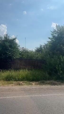 The video shows a view from a vehicle moving along a road on a sunny day. The road is paved with dirt and gravel. To the left of the road, there is a grassy area and a fence. Beyond the fence are trees and houses. The sky is blue with scattered white clouds. The scene appears to be in a rural or suburban area.