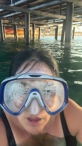 A woman wearing a snorkeling mask and a black swimsuit is in the water. She is looking at the camera and opening her mouth. The water is clear and green, and there are wooden pilings in the background. The sun is shining, and there are some boats in the distance. The woman submerges briefly before surfacing again.