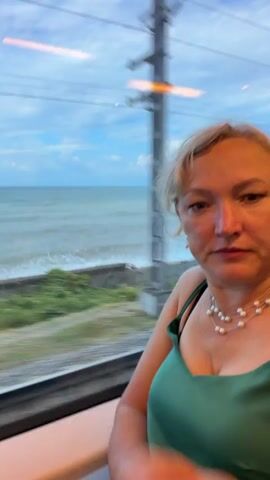 A woman with blonde hair and pearl jewelry, wearing a green dress, is seated by a window on a train. She is looking at the camera and smiling. Outside the window, the ocean and a cloudy sky are visible. The landscape is blurred due to the train's movement.