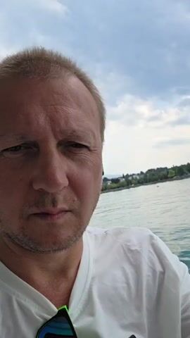 A man with blonde hair and a white t-shirt is taking a selfie on a boat. He has earbuds in both ears and sunglasses hanging from his t-shirt. The water is a bright turquoise color. In the background, there is a shoreline with trees and buildings. The sky is cloudy with some blue patches visible. The man appears to be looking directly at the camera.