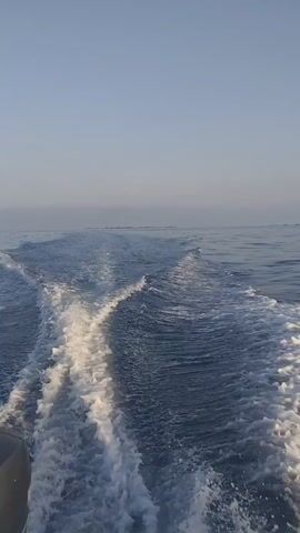 The video opens with a view of a boat's wake on calm blue water under a clear sky. The camera then pans up to show a fishing rod with its line trailing in the water as the boat moves forward. The camera continues to follow the fishing rod and the boat's wake. Finally, the camera returns to a shot of the wake on the water.
