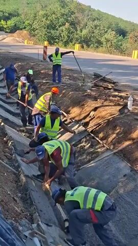 A group of construction workers are digging a trench. They are all wearing high-visibility vests. Some are using shovels, while others are using their hands to move dirt. The trench appears to be lined with concrete. There are trees and vegetation in the background, suggesting an outdoor environment. The weather appears to be sunny.