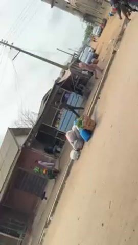 The video shows a street scene in what appears to be a small town. A large, weathered building with two towers, likely a mosque, is visible in the background. Along the side of the street, there are small, makeshift shops or stalls. In front of one of these stalls, there are several bags and a basket filled with items. People are walking along the street, some dressed in traditional clothing. The street itself is unpaved. Overhead power lines are visible. The overall impression is one of a bustling but modest urban environment.