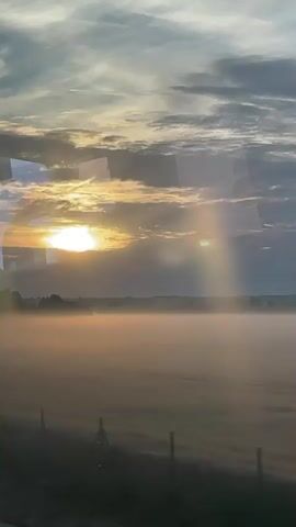 The video shows a rural landscape at sunrise, seen through a window. A field is covered in a layer of fog. The sun is rising behind clouds, casting light rays through them. A line of trees is visible on the horizon. A fence runs along the foreground of the field. The view is slightly obscured by reflections on the window.