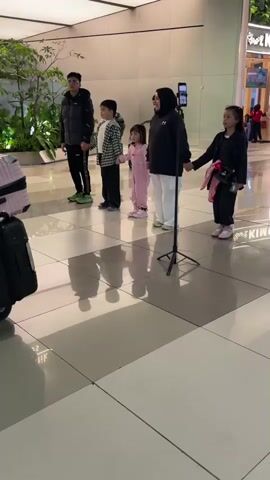 A group of five people, including two adults and three children, are standing in a line, holding hands. The group is facing a smartphone on a tripod, suggesting they are filming a video. The adults are positioned at either end of the line, with the children in the middle. The younger child, wearing a pink outfit, is on the left, and the older child, wearing a black outfit, is on the right. The man on the far left is wearing a black puffer jacket and black pants, while the woman in the middle is wearing a black hoodie and white pants. The background shows a modern, well-lit interior with large tiled floors and some greenery. The overall scene appears to be a public indoor space, possibly an airport or a shopping mall.