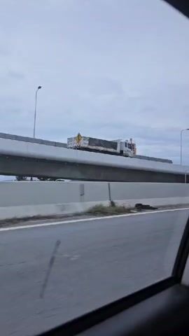 A truck is driving on an elevated highway. A camera mounted on a moving vehicle captures the scene from a low angle, looking up at the truck. The truck is white with a black tarp covering its cargo. It is moving from left to right across the frame. The truck is on an elevated section of a highway, with a concrete barrier separating it from the lower road below. The sky is overcast and cloudy. There are some green plants growing along the edge of the lower road. A yellow road sign indicating a merging lane is visible.