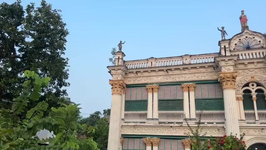 A slow-motion panning shot captures the ornate facade of a white, multi-story building with intricate architectural details. The building features rows of green-shuttered windows, decorative columns, and statues of small figures positioned on the roofline. In the foreground, lush green foliage and trees frame the view, with a decorative green metal archway and fence to the right. The sky above is a clear, pale blue.