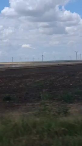 The video shows a wide shot of a rural landscape with a clear blue sky filled with puffy white clouds. In the middle ground, a field of wind turbines are visible stretching across the horizon. The foreground consists of a grassy field. There are no people or animals visible in the video.