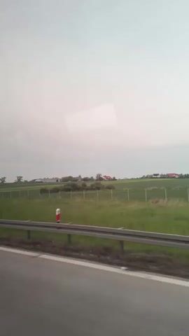 The video depicts a view from a vehicle traveling on a road. A metal guardrail runs along the side of the road, separating it from a green field. Beyond the field, there are some scattered buildings and trees. The sky is overcast and grey. As the vehicle moves forward, roadside signs appear, displaying text in a language other than English. The general environment seems to be rural.
