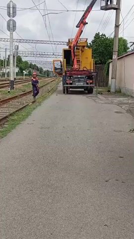 The video shows three workers repairing electrical lines on a railway. One worker stands on the ground near the tracks, while two others are elevated in a cherry picker attached to a yellow and black truck. The truck is parked on a narrow road adjacent to the railway. Above the cherry picker, the electrical lines extend across the tracks. Behind the road is a house with a fence and foliage. The sky is overcast.