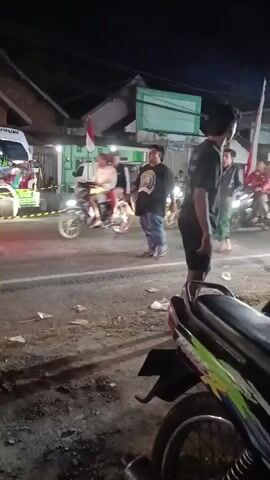 The video shows a nighttime street scene with several people and motorcycles. In the foreground, a motorcycle is parked, partially visible. Across the street, there are buildings and streetlights. Several motorcycles are in motion, with riders on them. One person, a male, is standing in the middle of the road, facing away from the camera, wearing a t-shirt with text on the back. Another male is walking across the street. There are flags visible, one red and white. The ground is paved and appears somewhat dirty with some litter. The lighting suggests it is dark outside, with artificial lights illuminating the area.