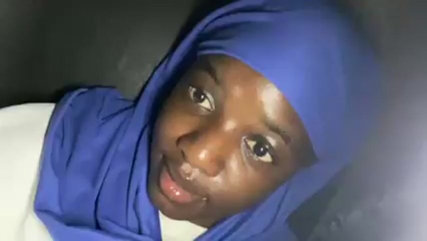 A close up video shows a person wearing a blue headscarf and white shirt, looking directly at the camera. The person's face appears shiny, and the background is dark. The person has dark skin and appears to be in a dimly lit environment.