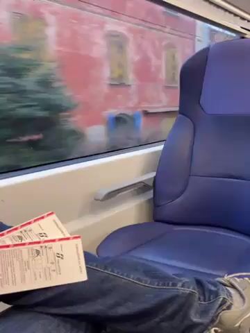 A person is sitting on a train, looking out the window at passing buildings. Three train tickets are resting on their lap. The seat is blue and the window frame is white. The person is wearing jeans and grey shoes. The train appears to be moving at a moderate speed. The weather outside is overcast.