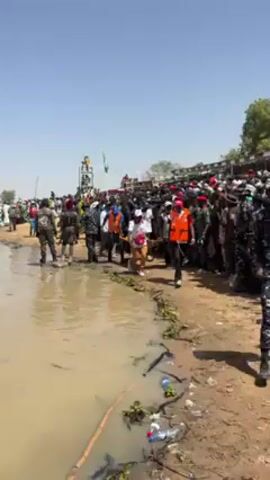 The video shows a crowded scene near a body of water, possibly a river or lake. People are gathered, including security personnel and medical personnel. Some individuals are wearing orange safety vests and others have white shirts with a red cross emblem, indicating a medical or first aid role. The medical personnel are carrying a person on a stretcher through the crowd. There is litter on the ground, including plastic bottles. In the background, there is a raised structure with people on top of it, possibly for observation or event staging. The weather appears sunny and clear. The scene suggests a public event or gathering where medical support is needed.
