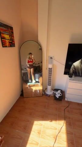A person with curly blonde hair, wearing a red sports bra and brown shorts, stands on a scale in front of a large, arched mirror. The person holds a smartphone in their right hand, taking a selfie, and flashes a peace sign with their left hand. White sneakers are worn. The mirror reflects a bedroom scene with a bed, a desk with items on it, and a window with string lights.

In the background, a tall, white tower fan stands next to a power strip with multiple plugs and a small black platform. The floor is tiled with terracotta-colored tiles. The room appears to be illuminated by natural light, suggesting it is daytime. The overall mood seems positive and lighthearted.

No visible text is present in the media file. The location context of Baan Bang Rak, Thailand, is not directly evident from the visual information within the frame.
