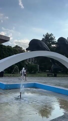 A video shows a fountain in the foreground with a sculpture of two figures hanging upside down in the background. A building with green ivy is visible behind the sculpture, and a tree is to the right. Several people are in the background: one person is sitting on a bench to the left, two people are walking near the fountain, and another person is standing near the fountain on the left. The sky is blue with some clouds.