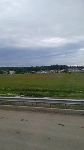 The video captures a view from a moving vehicle. In the foreground, there is a dirt road and a metal guardrail. Beyond the guardrail is a large, green field with scattered patches of white flowers. In the middle ground, there is a cluster of houses, some with visible roofs, situated at the edge of the field. A line of trees forms the background, and the sky is overcast with dark clouds. A tall, slender utility pole is visible in the left-center of the frame. The overall scene depicts a rural or semi-rural landscape under a cloudy sky.