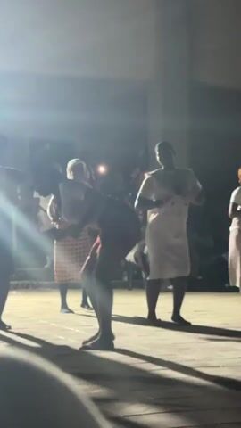 The video shows three women standing on a stage, illuminated by bright stage lights. They are all dressed in similar outfits: white shirts, knee-length skirts, and flat shoes. They appear to be performing a dance or movement routine, possibly clapping their hands in unison. The background is filled with a crowd of people, mostly sitting, watching the performance. The setting seems to be an indoor event, likely in a school or community hall, with white walls and large windows visible in the background. The video is somewhat blurry, particularly in the periphery, and the strong lighting creates significant glare and shadow.