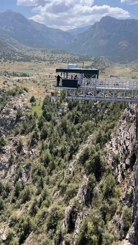 A person in red pants and a white shirt is bungee jumping from a platform. The person is suspended in mid-air by a bungee cord, with a vast, tree-covered canyon stretching out below. The platform is a modern, elevated structure with several people standing on it. The sky is blue with some clouds, and the sun is shining, creating a bright, clear scene. The overall impression is one of adventure and thrill-seeking in a dramatic natural landscape.