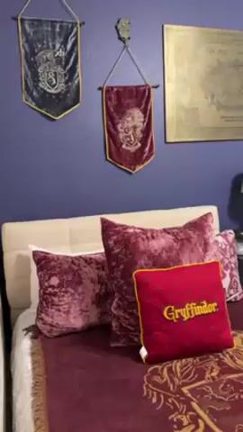 The video shows a bedroom with two twin beds decorated in a Harry Potter theme. The walls are painted a dark blue. Above the beds are banners with the logos of the Gryffindor and Ravenclaw Hogwarts houses. The bed on the left has a red and gold Gryffindor themed blanket and pillows, while the bed on the right has a blue Ravenclaw themed blanket and pillows. Each bed has a white headboard and a black nightstand with a lamp and various Harry Potter themed decorations. There is also a framed picture hanging on the wall.