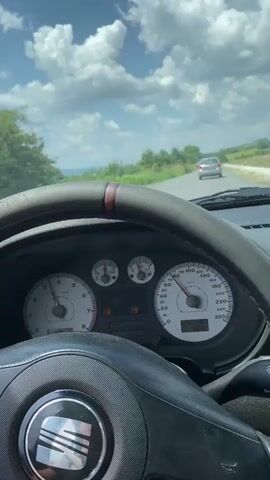The video shows the interior of a car driving on a road. The dashboard is visible, including the speedometer which reads approximately 100 km/h. The steering wheel is also visible. Outside the car, there is a landscape with hills, fields, and trees under a cloudy sky. A second car is seen ahead on the road. In one shot, a woman's face is visible near the window, and she is holding a cigarette.