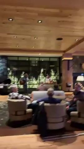 The video shows a well-lit hotel lobby with a live music performance. People are seated in comfortable armchairs and sofas, casually listening to the musicians. The lobby has a rustic-chic design with wooden ceiling panels, stone pillars, and large windows overlooking greenery. A bar area with illuminated shelves and bottles is visible in the background. The camera briefly pans around the lobby before moving towards a hallway with dark blue walls and a TV screen.