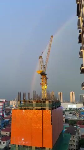 This media file captures a striking cityscape scene with a prominent construction site in the foreground. A tall yellow crane dominates the center of the frame, reaching towards a dramatic sky. The construction site itself is partially wrapped in bright orange netting, with scaffolding and rebar visible at the upper levels. Several figures, presumably construction workers, are present on the platform of the building under construction.

In the background, a soft rainbow arches across a grey, overcast sky, suggesting recent or ongoing rain. This natural phenomenon provides a beautiful contrast to the industrial elements of the construction site. Beyond the construction, a line of modern, tall buildings can be seen, indicating an urban environment. The lighting suggests it is either early morning or late afternoon, with the sky having a soft, diffused light.

The overall mood is one of dynamic contrasts: the raw, unfinished nature of construction against the ephemeral beauty of a rainbow, and the stark urban landscape softened by atmospheric conditions. The setting is clearly an urban area, specifically identified as Khan Mean Chey, Cambodia, given the presence of the construction and the architectural style of the background buildings. No visible text is present in the media file.