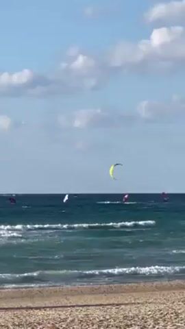 The scene is a beach on a partly cloudy day, with waves gently lapping the shore. Several individuals are kitesurfing in the sea, their colorful kites punctuating the horizon. A yellow kite stands out prominently in the center. The beach itself appears to be composed of small pebbles.