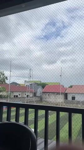 The video pans across a view from a balcony, looking out over a synthetic turf field marked with white lines, resembling a sports field. Beyond the field, there are several houses with varying roof colors, some red, some orange, and some with lighter tones. The sky is overcast with clouds. The balcony has a black railing and is covered by a black mesh netting. The video also briefly shows a black chair on the balcony.