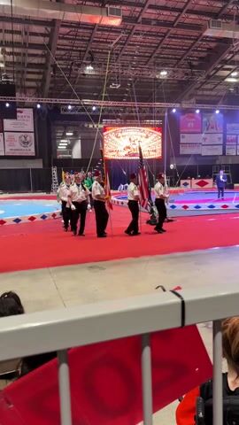 The video shows a group of men marching on a red carpet in what appears to be a circus arena. The men are wearing white shirts, black pants, and fezzes. They are carrying flags, including the American flag and a flag with a crescent and star symbol. In the background, there is a large LED screen displaying the words "Welcome to the Aladdin Shrine Circus". A man in a suit is standing on a platform nearby. There are advertisements for various businesses along the wall. The camera is positioned behind a metal railing, and a woman is visible in the foreground. The overall atmosphere is celebratory and patriotic.