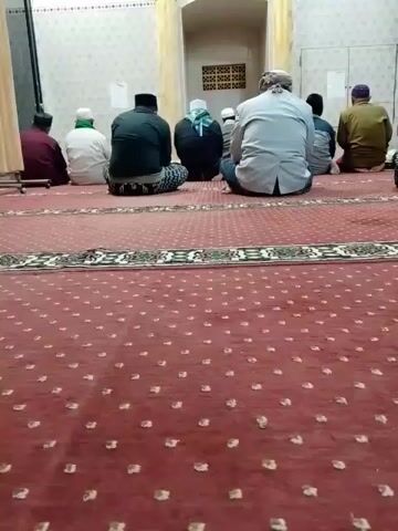 A group of men are sitting on the floor of a mosque facing a mihrab, a semicircular niche in the wall that indicates the direction of Mecca. The floor is covered with a red carpet with a repeating pattern of small white dots and a decorative border. The walls are tiled with a white and beige pattern. There are pillars on either side of the mihrab. A clock is visible on the wall above the mihrab. A ceiling fan is also visible. The men are dressed in traditional Islamic clothing, including kufis and robes. They appear to be engaged in prayer or contemplation.
