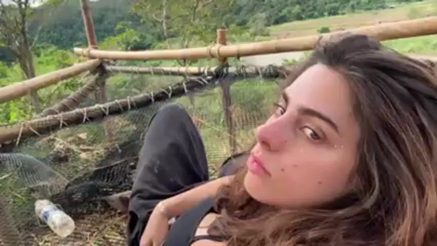 A woman with long brown hair is lying in a bamboo and net hammock. She is wearing a black top and black pants that appear to be dirty. The hammock is built overlooking a lush green valley with trees. A plastic water bottle lies near her feet in the hammock. The camera is angled above the woman, looking down at her. The surrounding environment appears to be a natural setting. The video is taken outdoors during the day.