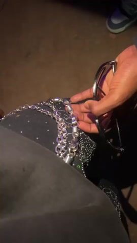 The video shows a close-up view of a person's hands holding sunglasses and a silver chain. The person is wearing black pants with some sort of sparkly embellishments on the left leg. The background is dark and appears to be outdoors on a textured surface. The person's fingers are manipulating the chain and sunglasses.