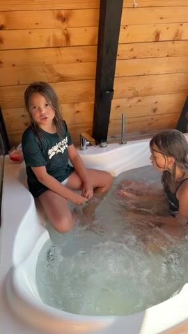 Two young girls are sitting in a hot tub. The girl on the left, with long brown hair, wears a dark green t-shirt and white shorts. She is looking at the camera with a neutral expression. The girl on the right, also with brown hair, wears a black swimsuit and is looking towards the camera with a slight smile. The hot tub is white and filled with bubbling water. The setting appears to be an outdoor space with wooden walls. In the background, beyond the hot tub, a scenic view of a village is visible, with houses, trees, a body of water, and mountains in the distance. The sunlight suggests it is daytime, likely late afternoon due to the warm lighting. The overall atmosphere seems relaxed and pleasant, with the girls enjoying their time in the hot tub. No text is visible in the media.