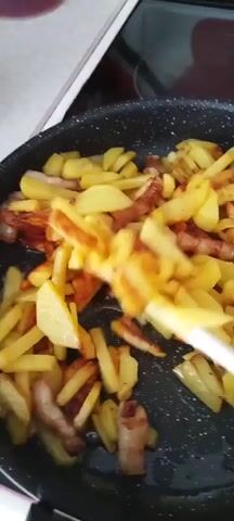 A close-up, overhead shot shows french fries and bacon being fried in a pan. A white spatula is visible on the right side of the frame, stirring the food. The potatoes are cut into irregular shapes and are golden brown, indicating they are close to being fully cooked. Pieces of bacon, some crispy and some still soft, are mixed in with the fries. The pan has a dark, speckled non-stick surface. The background is blurred but appears to be a kitchen environment.