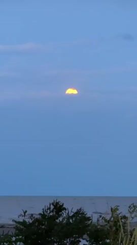 The video shows a yellow moon peeking from behind clouds in a blue sky over a body of water. In the foreground, there is some foliage. The water appears calm and the overall scene is serene. The moon is nearly full.